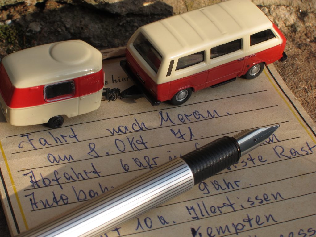 Nahaufnahme Zettel mit Reisenotizen, ein quer liegender Füller und darauf ein Spielzeugmodell eines T4 (Bully, VW Bus) mit 50er Jahre Wohnanhänger, beides in creme-rot Farben