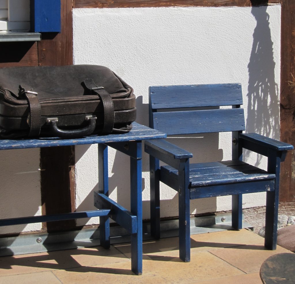 Ein kleiner blauer Tisch vor einem mutmaßlichen Fachwerkhaus, von dem nur ein kleiner Wandausschnitt zu sehen ist. Auf dem blauen Tisch liegt ein alter, kleiner schwarzer Koffer. Er ist geschlossen. Von Ansichtsseite rechts steht ein kleiner, leerer blauer Stuhl.