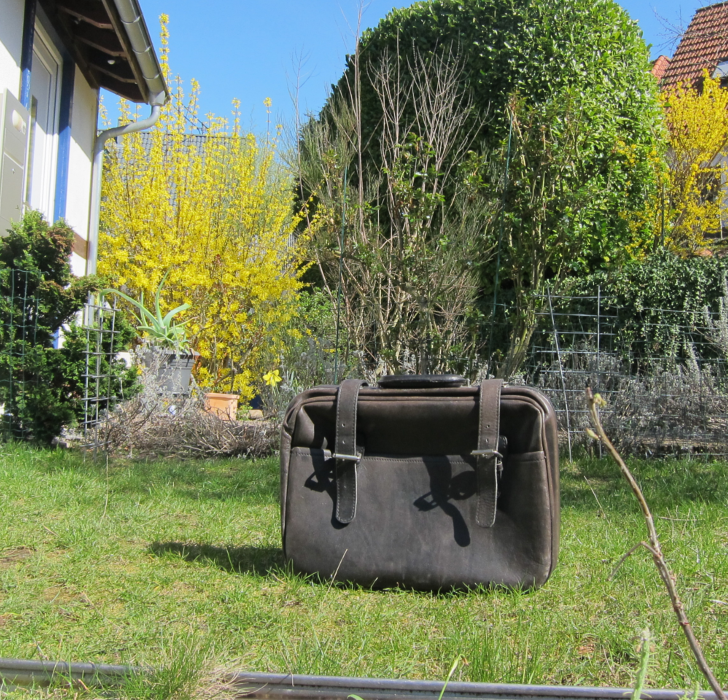 Kreisrundes Bild. In einem Frühlingsgarten steht ein kleiner schwarzer Koffer auf dem Gras. Im Hintergrund blüht eine Forsythie gelb.