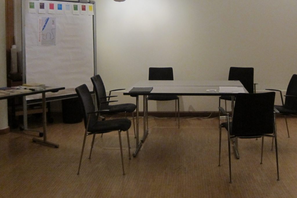 Raum in dem ein Tisch steht mit ein paar Papieren drauf. 6 freie schwarze Stühle stehen drumherum. Links ein Flipchart, an dem das Poster zur Schreibchallenge befestigt ist, schräg davor steht der Büchertisch.
